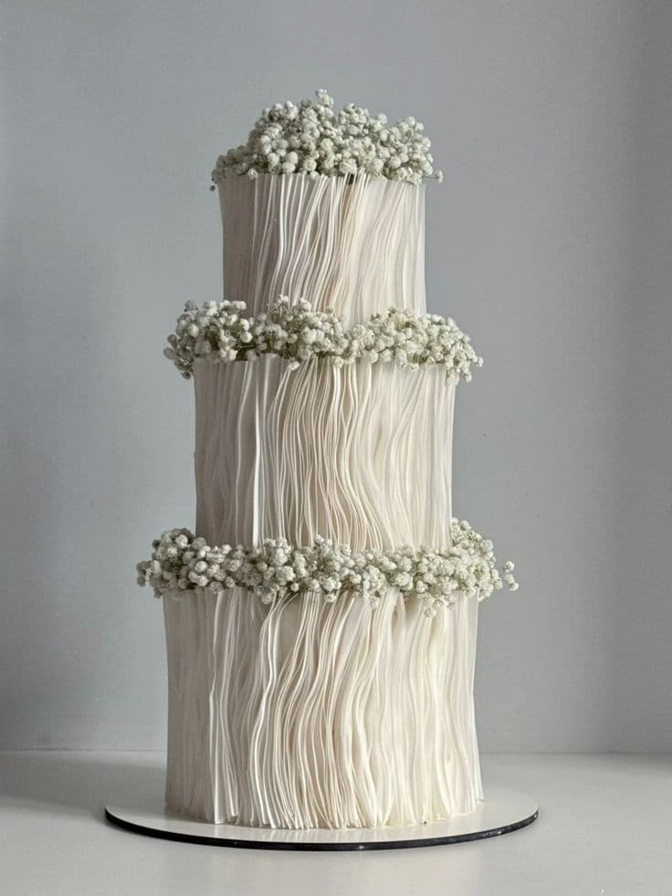 A three-tiered white cake featuring handcrafted vertical pleats and rustic rings of fresh baby's breath flowers between each layer and on top. A tall, minimalist three-tiered custom cake. The white frosting is textured with deep, handcrafted vertical waves. Delicate white baby's breath flowers form a snug ring between the first and second tiers, the second and third tiers, and crown the top of the cake.