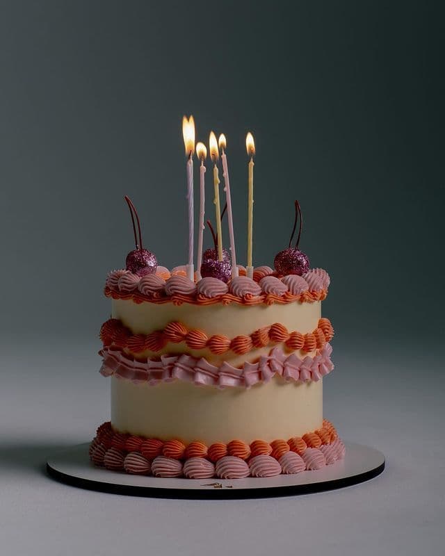 A tall, two-tiered custom white cake. The top tier is finished with perfectly smooth frosting and sharp edges, while the entire bottom tier is heavily textured with intricate, overlapping frills resembling draped fabric or petals. A beautiful, realistic cascade of handcrafted white sugar roses and delicate light-green leaves flows diagonally down the side of the cake. Handcrafted elegant white wedding cake by PlateStory in Chennai, featuring a smooth top tier, heavily textured ruffled fondant base, and realistic white sugar flowers with green leaves.