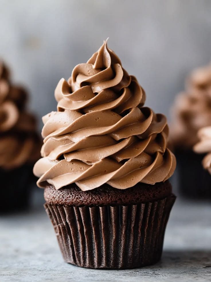 A close-up view of a freshly baked chocolate cupcake in a dark paper liner. The top of the cake is generously frosted with a tall, smooth, and glossy swirl of dense 46.5% dark chocolate ganache, highlighting its rich, velvety texture. Handcrafted dark chocolate cupcake by PlateStory in Chennai, featuring a moist cocoa sponge and a glossy swirl of premium 46.5% dark chocolate ganache.