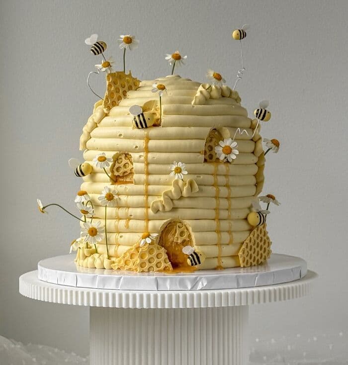 A custom cake sculpted into the shape of a domed beehive using thick horizontal bands of yellow frosting. The cake is decorated with handcrafted yellow and black bees, some attached directly and others flying on thin wires. White daisy flowers with yellow centers, textured honeycomb pieces, and realistic honey drips complete the design.