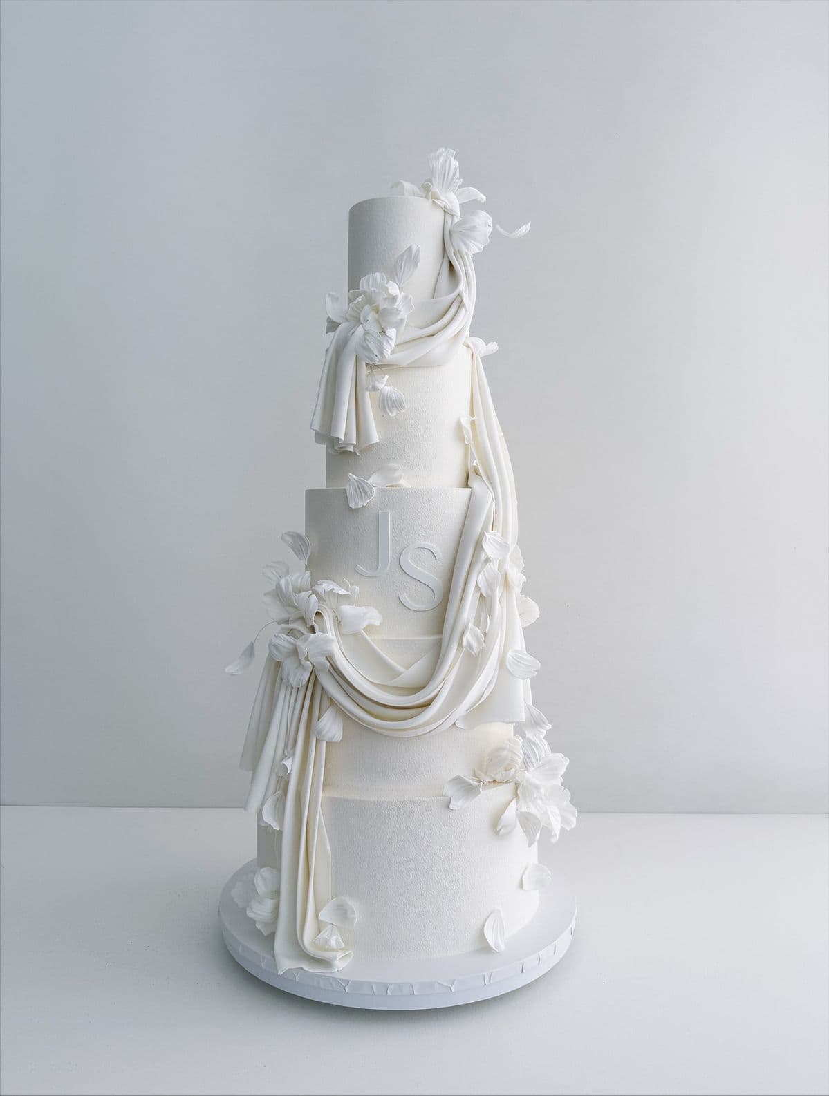 A towering five-tier white custom cake on a white surface. The tiers have a subtle, stippled texture. A piece of realistic, white fondant fabric is sculpted to look like it is draping elegantly down from the top tier to the bottom, intertwined with delicate white sugar flowers and loose petals. The middle tier features a large, clean, recessed monogram of the letters J and S. A tall, five-tiered white cake featuring textured icing, draped white fondant fabric, white flowers, and a 'JS' monogram.
